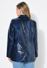 Niebieska sztuczna skóra w motyw węża, z błyszczącym wykończeniem, z klapami oraz pojedynczym rozcięciem z tyłu, w połączeniu z jasnymi dżinsami.