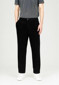 Mann wearing sorte plisserte bukser med sidelommer, en falmet mørkegrå kortermet skjorte og hvite joggesko på en ensfarget bakgrunn.