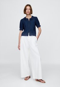 Mörkblå denimblus med rundad krage, kort puffärm och spetskant; kombinerad med vita vida byxor och bruna sandaler.