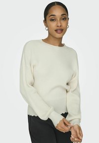 Maglione lavorato a maglia in bianco sporco, caratterizzato da una texture a coste e un orlo ondulato. Il design include maniche lunghe e un colletto rotondo.