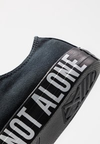 Chaussure en toile noire avec semelle texturée noire, présentant le texte « NOT ALONE » en lettres blanches audacieuses sur le côté. Design simple avec couture discrète.