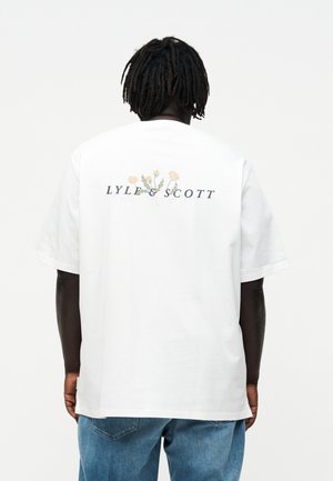 Personne avec des dreadlocks portant un t-shirt blanc avec un motif floral et le texte "LYLE & SCOTT" dans le dos, associé à un jean bleu.
