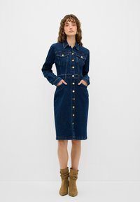 Denimmekko, jossa on napitettava etuosa, pitkät hihat, rintataskut ja sivutaskut. Tumma sininen väri, vyötä ympäröivä malli ja polveen ulottuva helma.