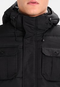 Svart vadderad jacka med hög krage, två framfickor, dragsko på huvan och svarta knappar. Har en slät yta och minimalt med sömmar.