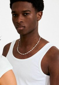 Valge soonikkoega tanktop, hõbedast ketti värritatud kaelakee, tume nahatoon, lühike juukselõikus, ühevärviline taust.