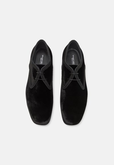 Chaussures en velours noir avec un profil bas, bout pointu, design à lacets et couture complexe le long des bords. Texture lisse, détails minimaux.