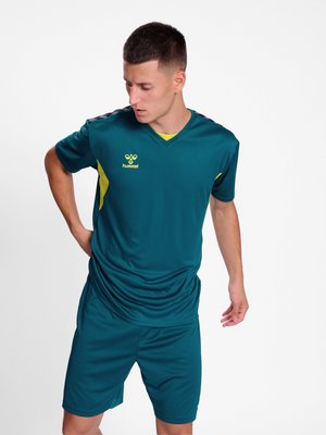 Młody mężczyzna w turkusowej odzieży sportowej z żółtymi akcentami, patrzący w dół, z jedną ręką na biodrze, na jednolitym białym tle.