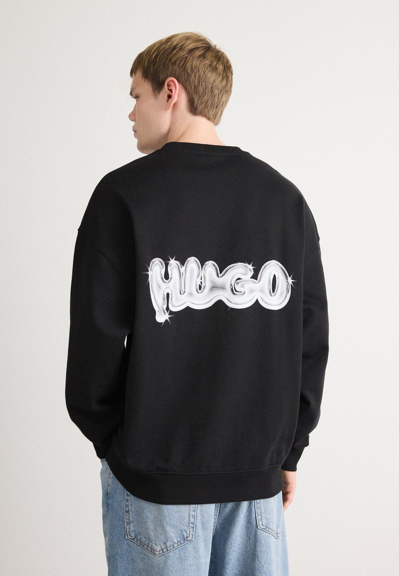 Sudadera negra con un gran logo blanco estilizado "HUGO" en la parte posterior, con una textura suave y puños acanalados. Diseño de corte casual.