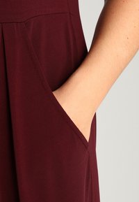 La mano destra inserita nella tasca laterale di un indumento color bordeaux, mostrando il tessuto liscio e i dettagli delle cuciture visibili.