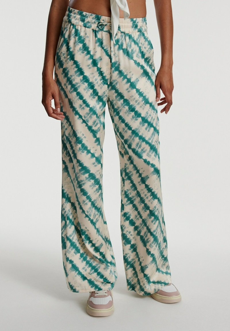 Personne portant un pantalon large tie-dye crème et turquoise, des baskets blanches et un crop top blanc, debout devant un fond uni.