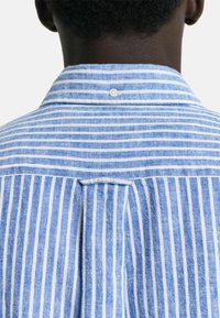 Camicia a righe blu e bianche con yoke posteriore, dettaglio dei bottoni e tessuto strutturato, con righe verticali di larghezze variabili.