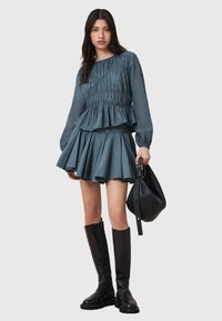 Blusa peplum turchese con texture arricciata, abbinata a una gonna turchese a campana. Il modello indossa stivali neri fino al ginocchio e tiene una borsa nera.