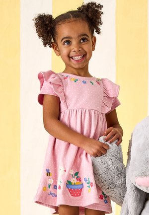 Noor tüdruk lokkis patsidega roosas kleidis hoiab kinni suure halli pehme elevandi käest triibulise kollakasvalge tausta ees.