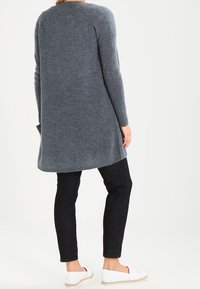 Grå stickad kofta med långa ärmar och rundad nederkant, i kombination med svarta smala byxor och vita slip-on skor. Mjuk textur.