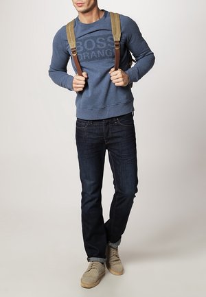 Modrá mikina se textem "BOSS ORANGE", tmavé džíny a světlé boty. Hnědé popruhy na batohu jsou viditelné.