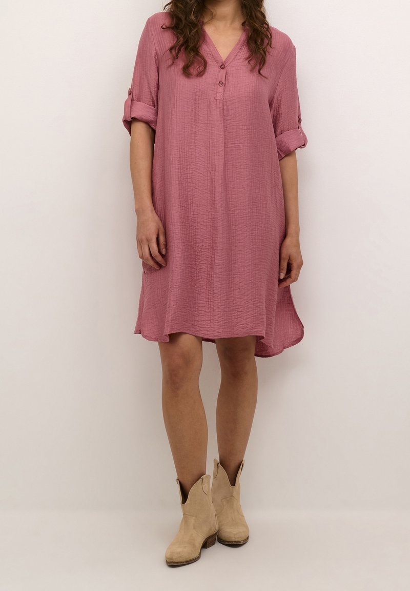 Robe rose à boutons avec un col en V, manches retroussées et tissu texturé. Conçue avec un ourlet asymétrique, associée à des bottines beige.