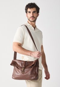 Brown leather messenger bag with a fold-over flap, metallic clasp, and a long strap, featuring a smooth texture and minimalist design.