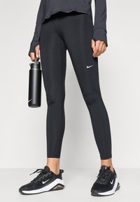 Leggings negre, dintr-un material aderent, cu o textură netedă și un logo Nike alb contrastant, asortate cu pantofi sport negri și o sticlă de apă neagră.
