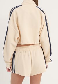 Chaqueta corta beige con textura de gofre, con rayas azul marino y blancas; pantalones cortos a juego con cintura elástica; ajuste relajado y silueta deportiva.