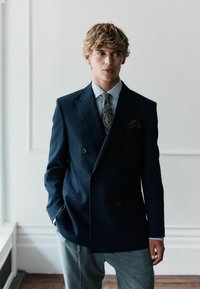 Tumes sinine topeltkinnitatud bleiser suuremate nööbidega, kombineeritud heleda triibulise särgi ja mustriga lipsuga. Hallid kohandatud püksid täiendavad välimust.