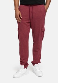 Burgunderfarbene Cargo-Jogginghosen mit einem Kordelzugbund, Seitentaschen und einem weichen, strukturierten Stoff, kombiniert mit weißen Sporthalbschuhen.