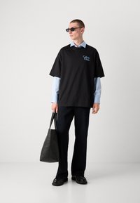 T-shirt en coton noire oversize avec un logo bleu, superposé à une chemise bleu clair. Pantalons sombres et sac en cuir noir. Lunettes de soleil claires.