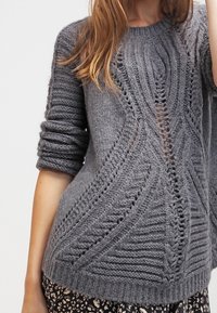 Gray knitted sweater featuring a textured pattern with ribs and holes, round neckline, and loose fit sleeves. Worn over patterned fabric.