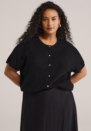 Kvinde med krøllet hår iført sølvfarvede creoler, en sort tekstureret kortærmet cardigan med knapper og et sort skørt, poserer foran beige baggrund.