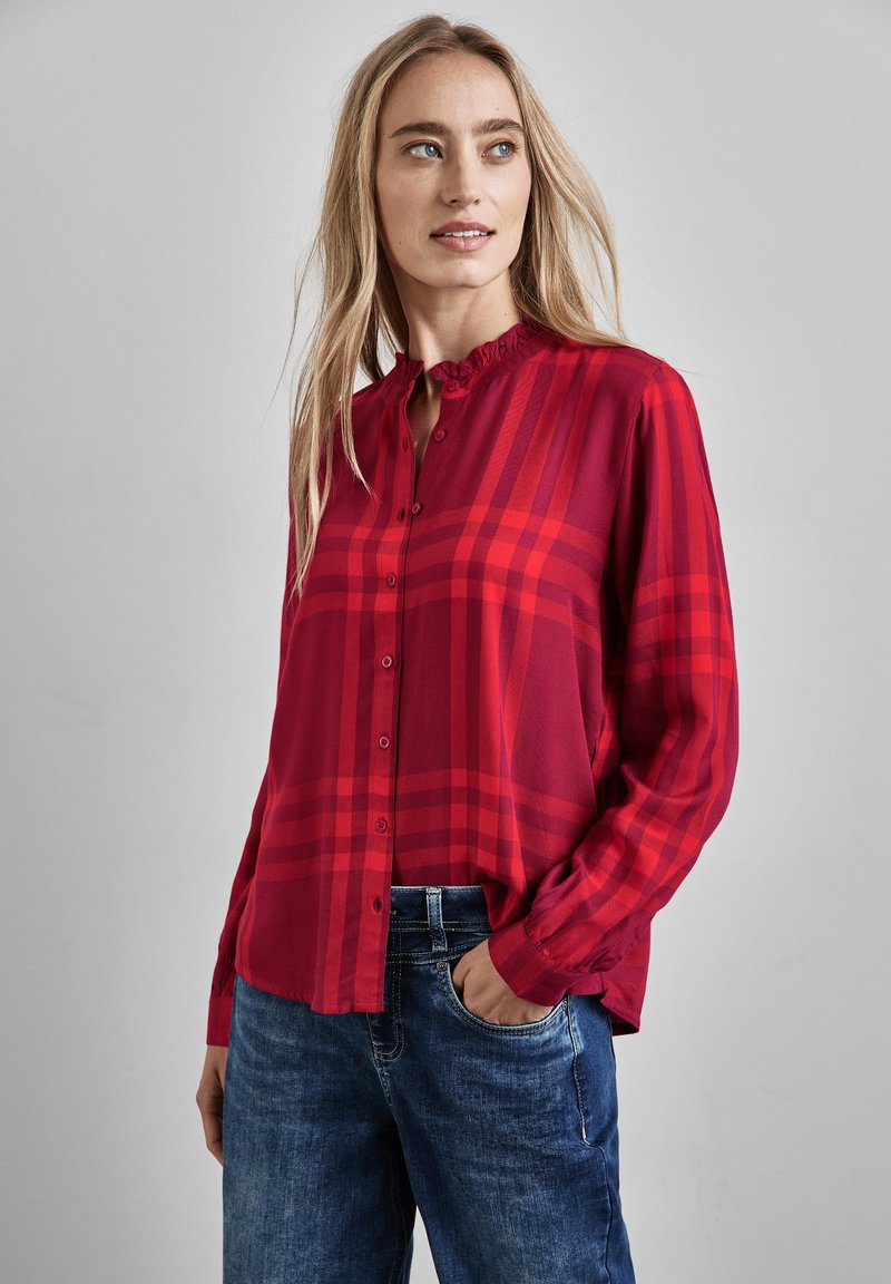 Camicia a quadri rossa con colletto arricciato, maniche lunghe e una vestibilità ampia. Indossata con jeans blu, presenta un tessuto testurizzato.