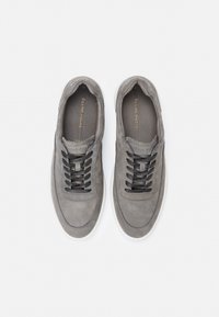 Szare zamszowe trampki z czarnymi sznurowadłami, zaokrąglonym noskiem, białą gumową podeszwą i wytłoczonym logo na języku. Gładka faktura i minimalistyczny design.