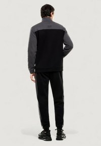 Chándal deportivo negro y gris con chaqueta de cremallera, con cuello alto y puños elásticos, combinado con joggers a juego y zapatillas.