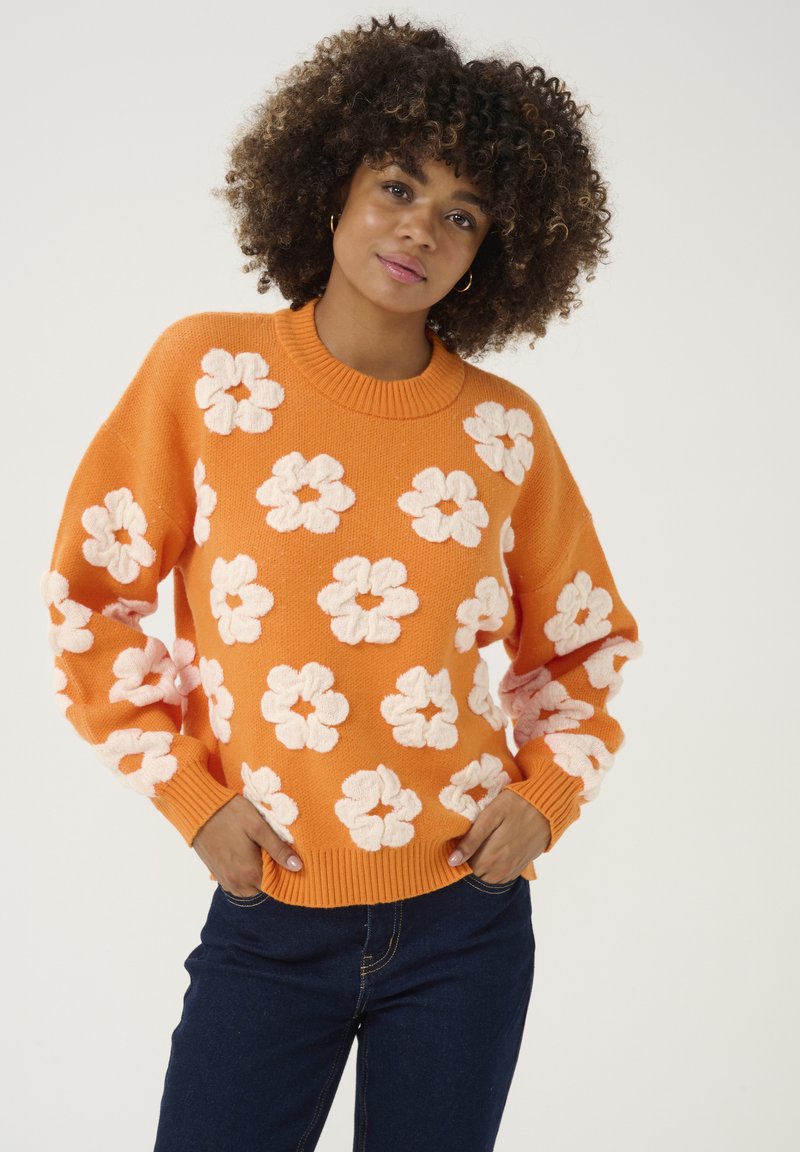 Félénk narancssárga pulóver texturált fehér virágmintával, bordás nyakrésszel és mandzsettákkal. Sötét farmernadrággal párosítva, laza szabású dizájn.