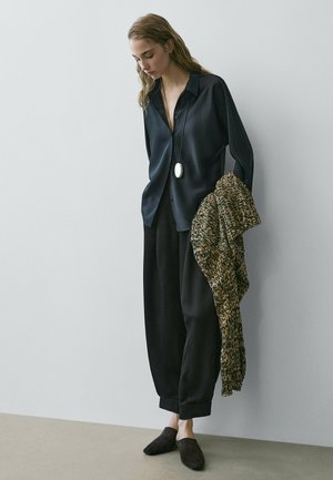 Vrouw draagt een losse zwarte blouse en broek, houdt een gestructureerde bruine en zwarte trui vast, staand tegen een effen lichte muur.