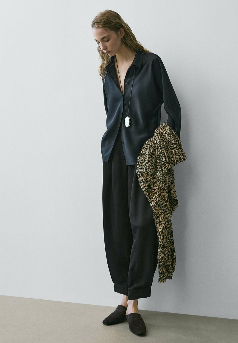 Femme portant une chemise noire ample et un pantalon, tenant un pull texturé marron et noir, debout devant un mur clair uni.