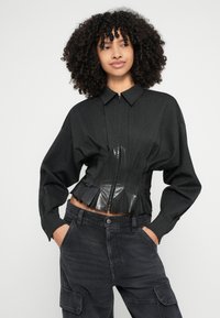 Giacca nera corta con un davanti strutturato, caratterizzata da un pannello in plastica nera lucida a contrasto e accenti plissettati. Abbinata a pantaloni cargo scuri.