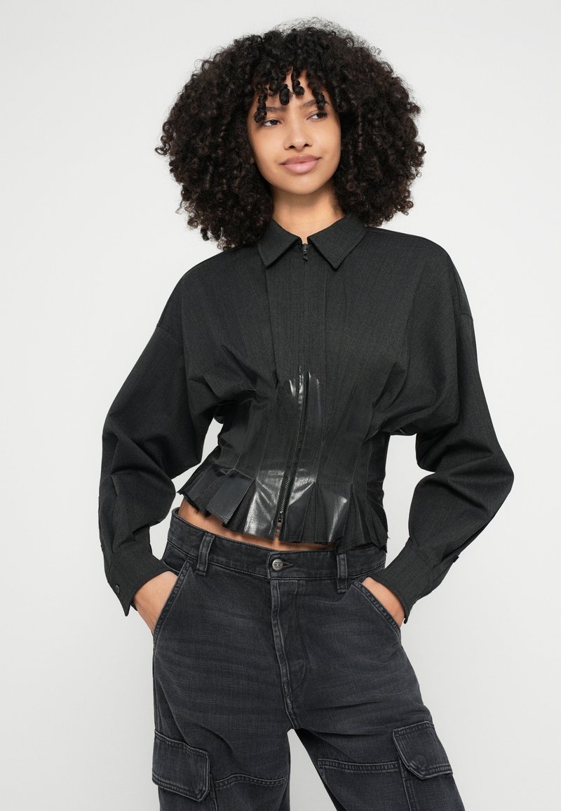 Schwarze, kurz geschnittene Jacke mit einer strukturierten Vorderseite, die ein kontrastierendes, glänzendes schwarzes Kunststoffpaneel und plissierte Details aufweist. Kombiniert mit dunklen Cargo-Hosen.