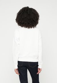 Felpa bianca con maniche lunghe, polsini e orlo a costine, vestibilità rilassata, abbinata a jeans in denim scuri, vista da dietro.