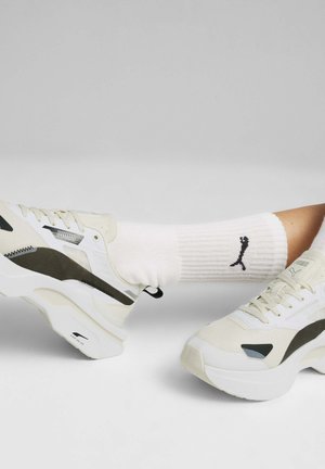 Zapatillas de deporte de color claro con malla texturizada y acentos sintéticos, que cuentan con una suela gruesa. Un calcetín blanco muestra un logo negro en el puño.