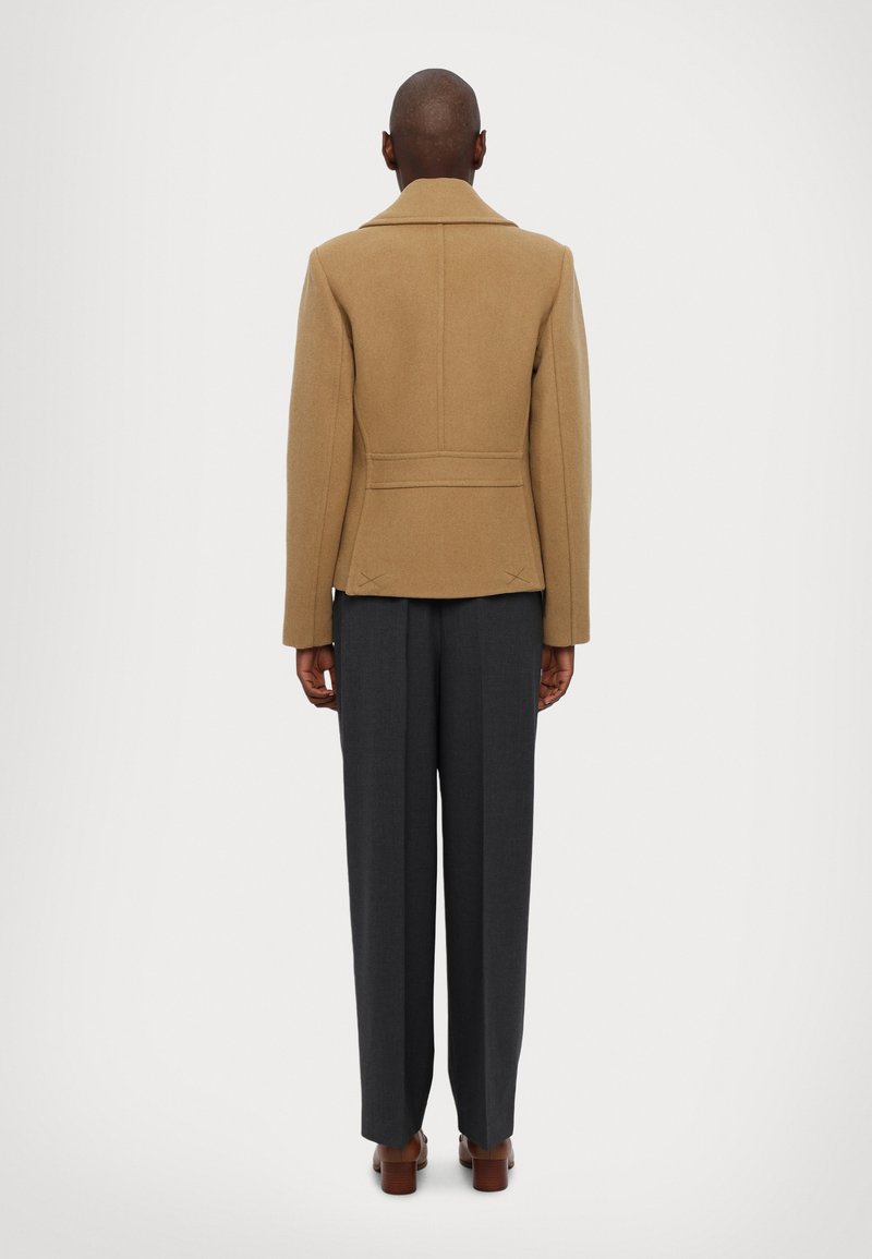 Kamelbraune Wollmischjacke mit schlanker Silhouette, Rückennähten und breitem Kragen, kombiniert mit dunkelgrauen, taillierten Hosen.