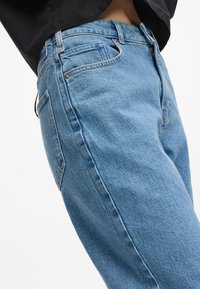 Svetlomodré džínsy s rovnými nohavicami, viditeľným šitím a malou modrou nášivkou pri páse. Štruktúrovaná tkanina s vreckami.