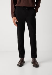 Persona vestida con pantalones negros de cordón, camisa de manga larga marrón, calcetines grises y mocasines de cuero marrón oscuro, de pie frente a un fondo liso.