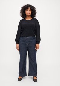 Black top with textured bodice and long, puffy, solid sleeves; paired with wide-leg dark denim jeans. Model stands in casual flats.