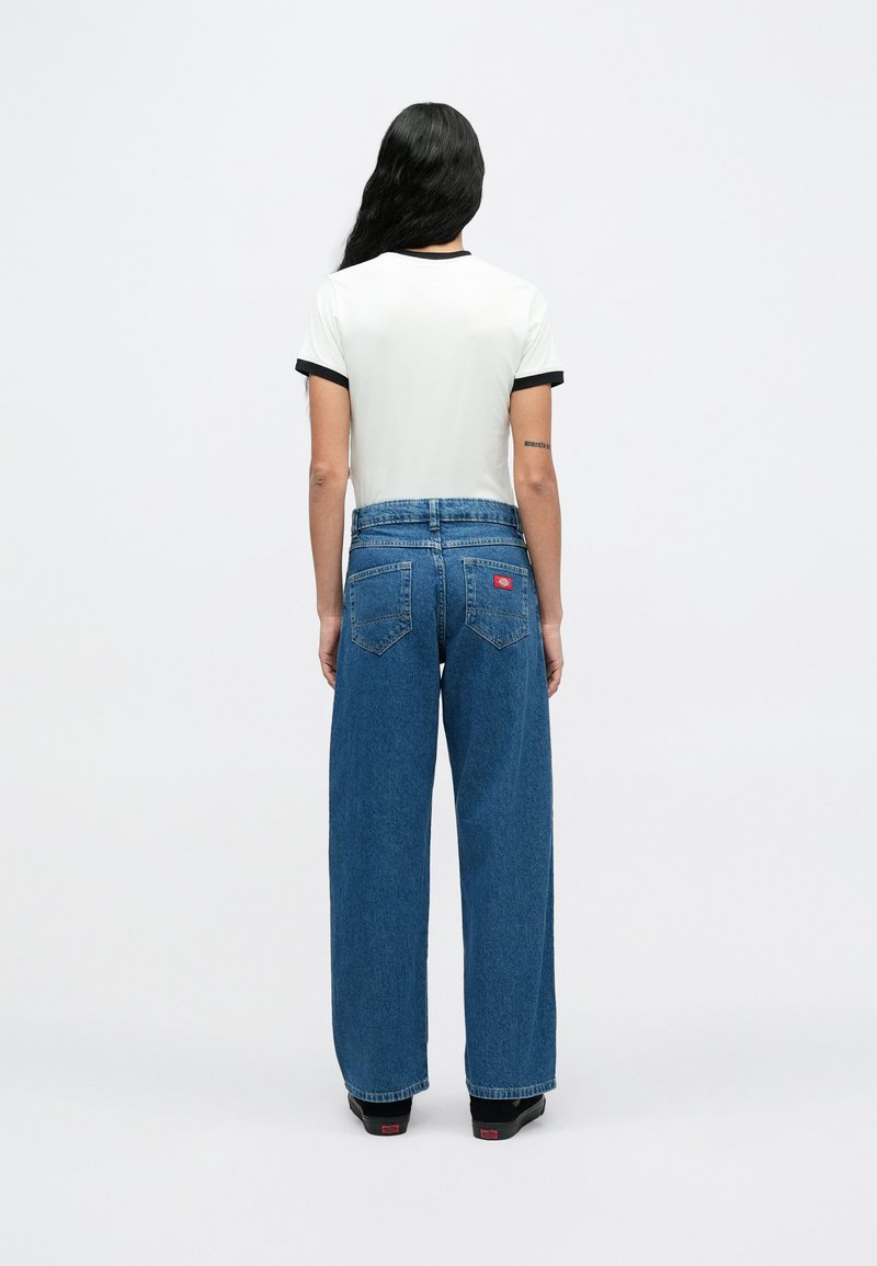 Femme aux longs cheveux noirs vue de dos, portant une chemise blanche à manches courtes avec bordure noire, un jean bleu à jambes larges et des chaussures noires.