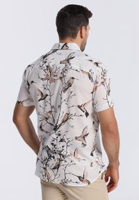 Chemise à manches courtes avec un fond blanc et un motif détaillé d'oiseaux et de branches dans des couleurs douces et atténuées. Col boutonné avec une coupe décontractée.