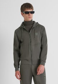 Felpa grigia con zip, dotata di tasche frontali, tessuto testurizzato, cordini regolabili e una toppa con logo. Abbinata a pantaloni coordinati e occhiali da sole.