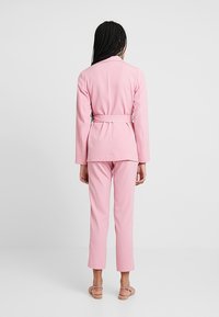 Femme aux cheveux tressés portant un blazer rose ceinturé, un pantalon assorti et des sandales, se tenant devant un fond clair uni.