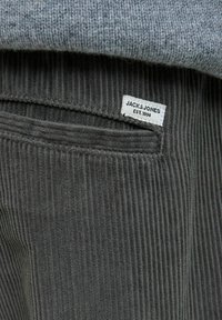 Sivkasta korduroj tkanina z navpičnimi rebri. Vsebuje majhno belo oznako z napisom "JACK & JONES EST. 1990" na pasu.