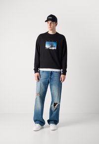 Felpa nera con stampa grafica, abbinata a jeans azzurri a gamba larga con strappi evidenti. Sneakers bianche e cappellino nero.