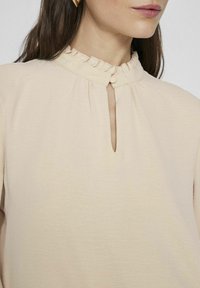 Krämsfärgad blus med volangkrage, nyckelhålshalsringning, mjuk textur och mjuka veck. Minimalistisk design med en neutral färg.