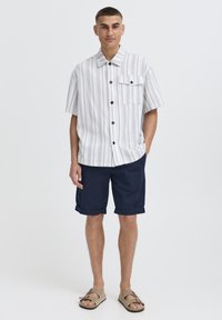 Cămașă cu mâneci scurte, cu dungi în alb, albastru și bej, cu nasturi, asortată cu pantaloni scurți bleumarin și sandale bej, având un buzunar la piept și manșete rulate.